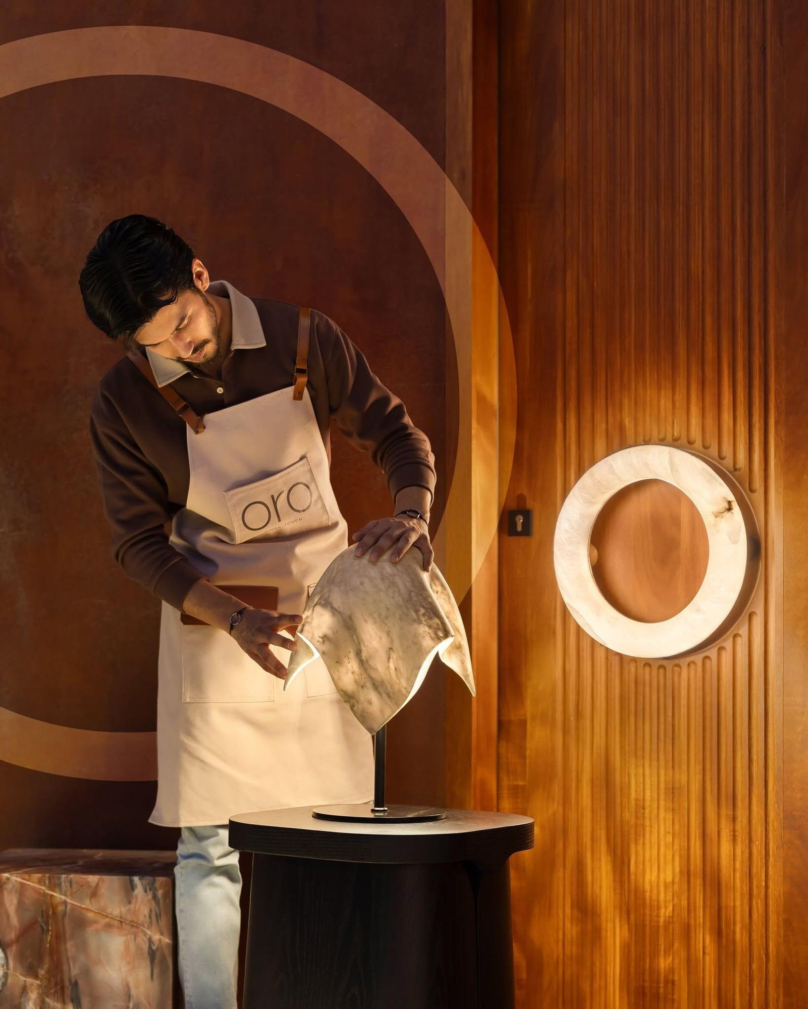 A maker in an oro apron finishing an alabaster light in the studio, an onyx ring glowing on the wall behind